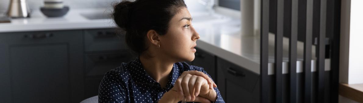 Vrouw met donker haar, zit in woonkamer en kijkt peinzend naar buiten door het raam