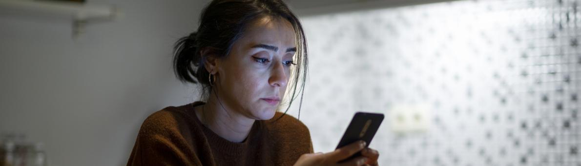 Vrouw zit aan tafel in haar keuken en kijkt peinzend op haar smartphone