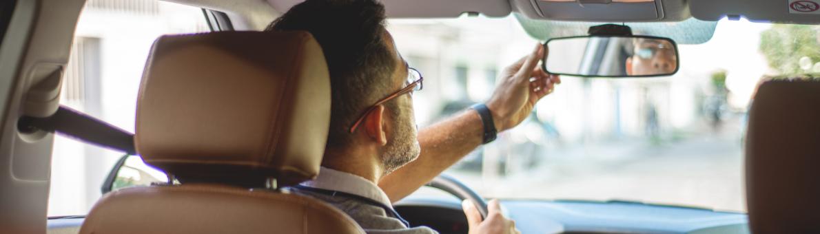 Man in auto op bestuurdersstoel van achteren gefotografeerd. Hij houdt zijn achteruitkijkspiegel vast en kijkt daar in.