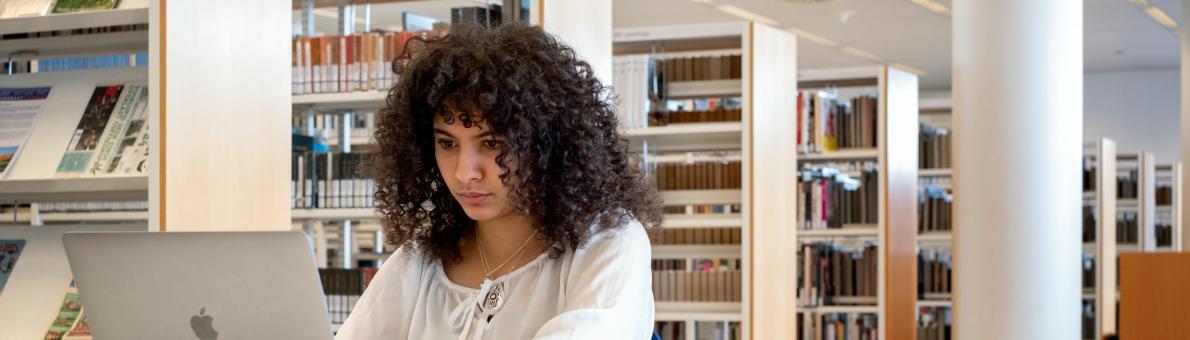 Jonge vrouw zit aan een tafeltje in een bibliotheek met haar laptop voor zich opengeklapt.