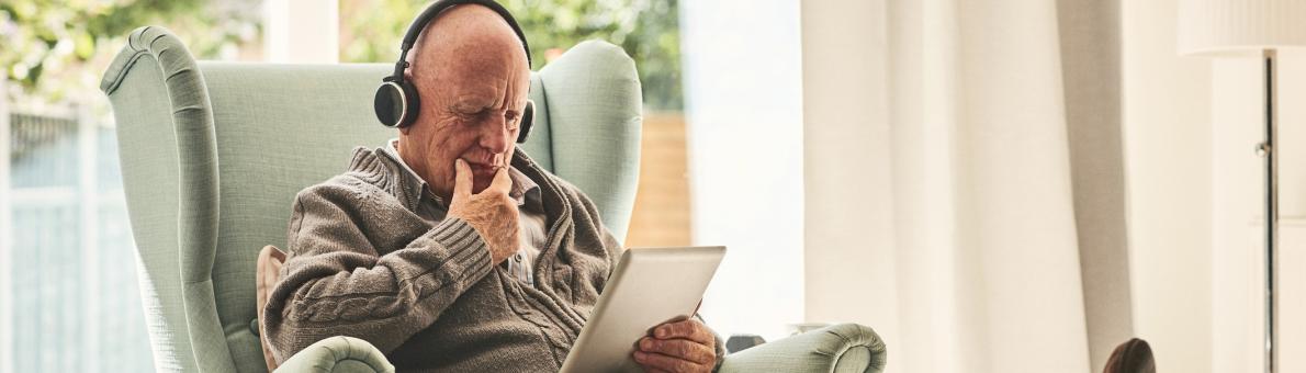 Oudere man zit op stoel met koptelefoon op zijn hoofd en tablet in zijn hand.