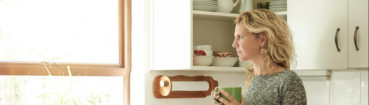 Een vrouw met blond krullend haar staat in haar keuken en kijkt uit het raam. In haar hand heeft ze een mok.