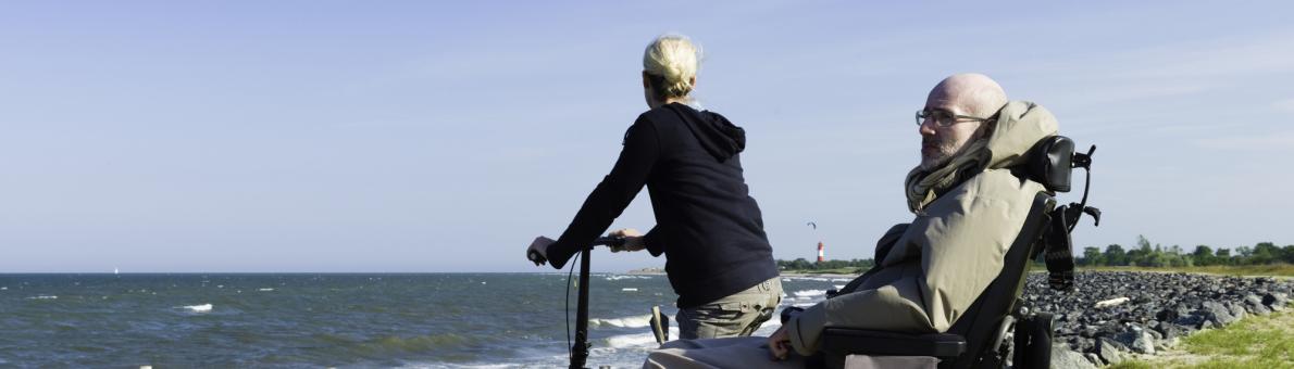 Een senior man zit in een scootmobiel. Naast hem staat een vrouw met een fiets. Ze staan op de rotsen dicht bij zee.