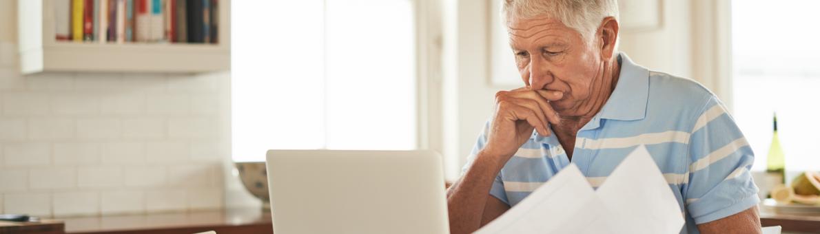 Een oudere man zit bezorgd achter zijn computer met papieren in zijn hand