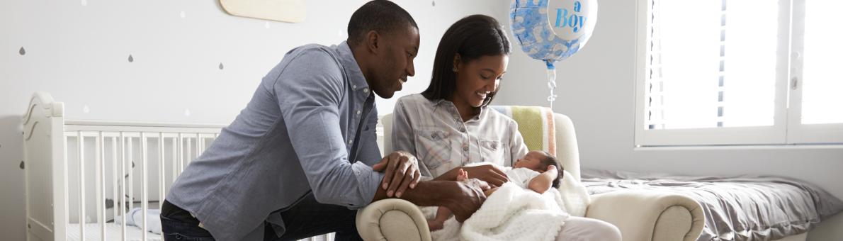 Vrouw zit met baby in haar armen op een stoel. Haar man zit geknield achter haar en kijkt glimlachend naar de baby.