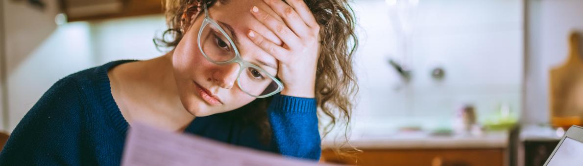 Een vrouw zit met papieren in haar hand aan tafel. Voor haar staat een laptop. Ze kijkt zorgelijk. Ze draag een bril en heeft een blauwe trui aan.