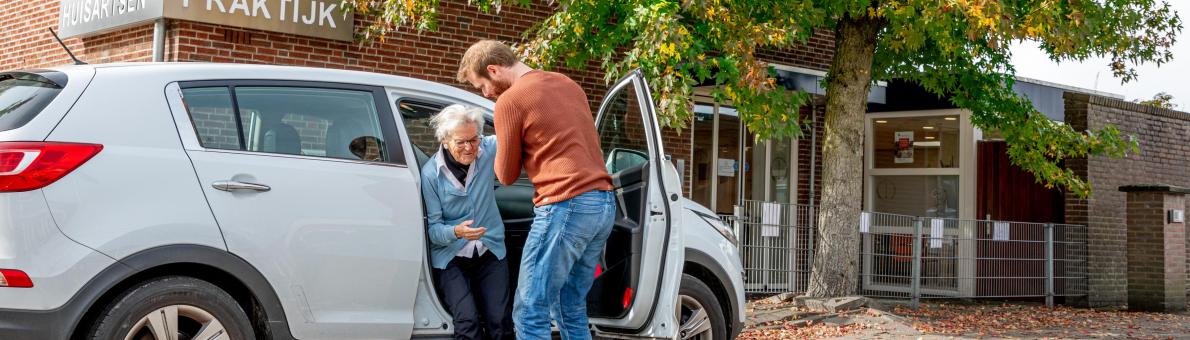 Mantelzorger helpt oudere vrouw uit de auto. Ze staan geparkeerd voor de huisartsenpraktijk.
