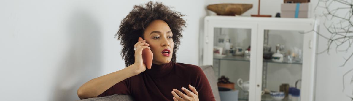 Een volwassen vrouw zit in kleermakerszit op de bank. Ze is aan het bellen.