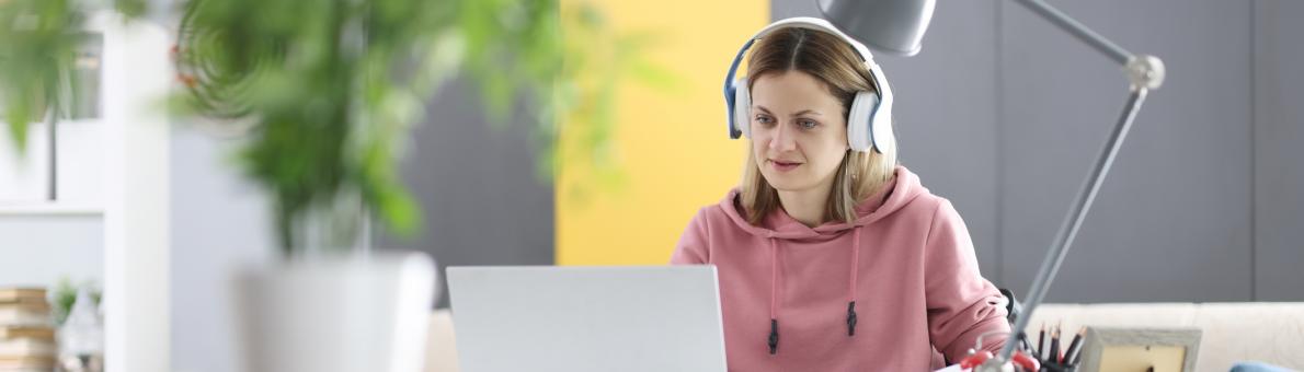 Een jongvolwassen vrouw in een rolstoel zit thuis achter haar laptop te werken of studeren. Ze draagt een roze trui en heeft een koptelefoon op.