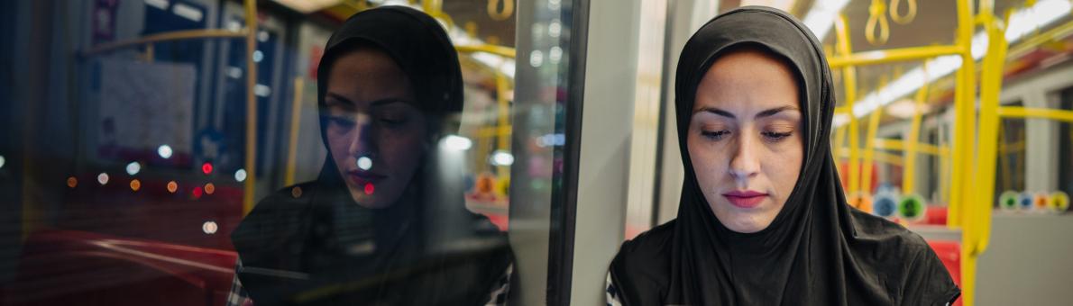 Een vrouw van middelbare leeftijd met hoofddoek en geblokte blouse zit met haar telefoon in haar hand in het openbaar vervoer.