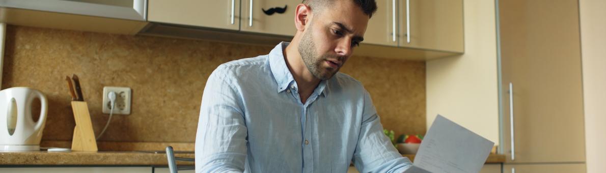 Een man van middelbare leeftijd met donker haar en een blauwe blouse zit aan zijn keukentafel. Hij leest een brief.