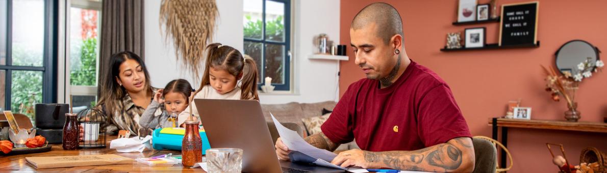Jong gezin van vader, moeder en twee jonge dochters regelt thuis zaken achter laptop. Het is een kleurrijke omgeving.