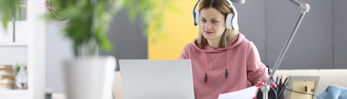 Jongvolwassen vrouw in rolstoel zit achter haar bureau. Ze heeft een koptelefoon op en kijkt naar haar laptop.