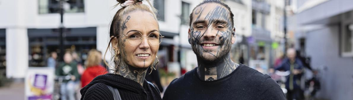 Jenny en William, een vrouw en man staan naast elkaar op straat. Ze zijn beiden zichtbaar getatoeëerd.