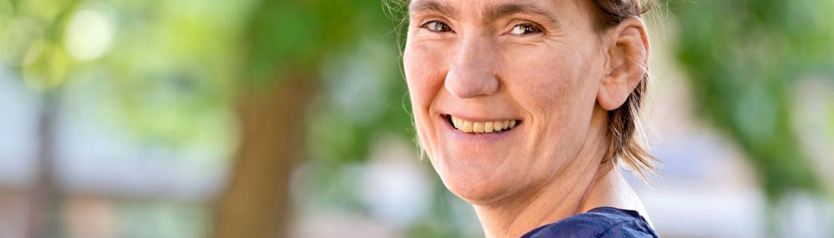 Lachende vrouw (Gudele Boland) met kort haar en blauw shirt. Op de achtergrond staat een boom