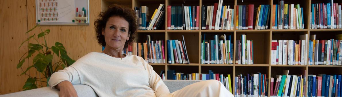 Vrouw met donkere krullen zit op de bank. Achter haar staat een boekenkast Vrouw met donkere krullen zit op de bank. Achter haar staat een boekenkast
