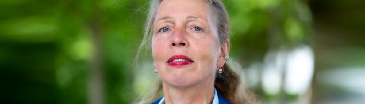 Vrouw (Janny Arends) staat in het park. Haar in een staart, rode lippenstift en draagt blauw jasje 