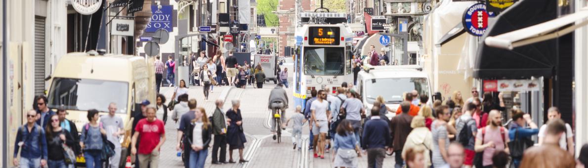 Een foto van een straat in Amsterdam. Je ziet mensen wandelen, tramrails, en aan beide kanten winkels.