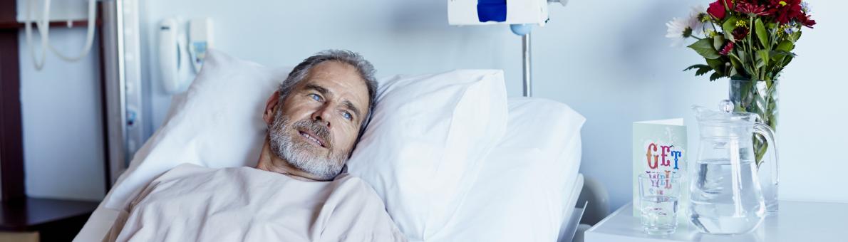 Man ligt in ziekenhuisbed. Hij heeft een baard en draagt een licht gekleurd shirt. Naast het bed staan bloemen.