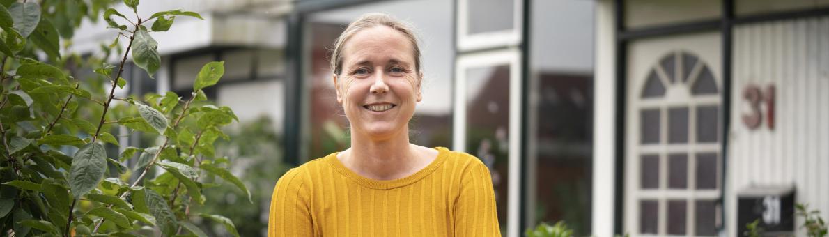 Vrouw met gele trui staat glimlachend voor haar huis