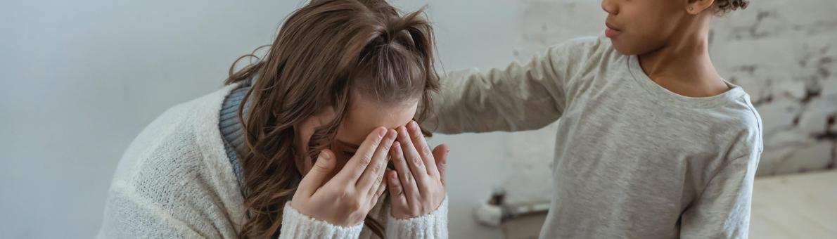 Jonge moeder zit voorover gebogen met handen voor gezicht, kindje troost haar