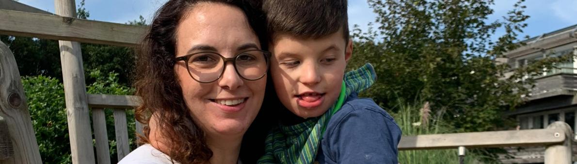 Vrouw met donkerbruin haar en bril kijkt lachend in de camera. Ze houdt haar gehandicapte zoon met donkerbruin haar in haar arm