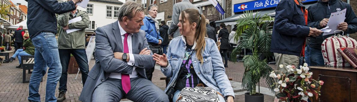 De Nationale ombudsman op bezoek in Zeeland. Hij is in gesprek met een vrouw met donkerbruin lang haar. Ze bevinden zich op een plein.