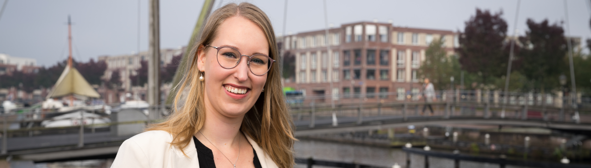 Vrouw (Michelle Koelewijn) heeft blond haar en draagt een bril. Ze staat buiten bij het water (haven)