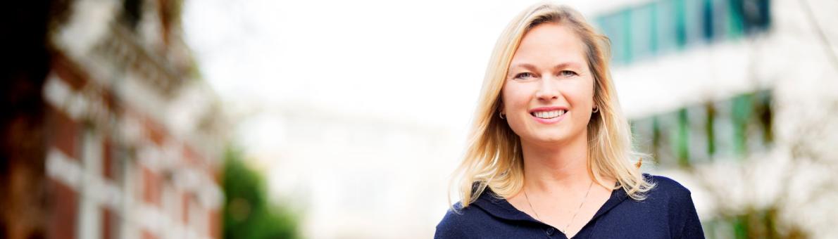 Blonde vrouw (Evalien van Toor Nationale ombudsman) kijkt lachend in de camera. Ze staat op straat en draagt een blauwe jurk