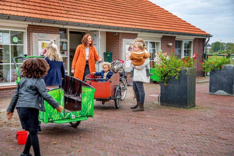 Vrouw in lange oranje jas haalt kinderen op bij de kinderopvang met een bakfiets