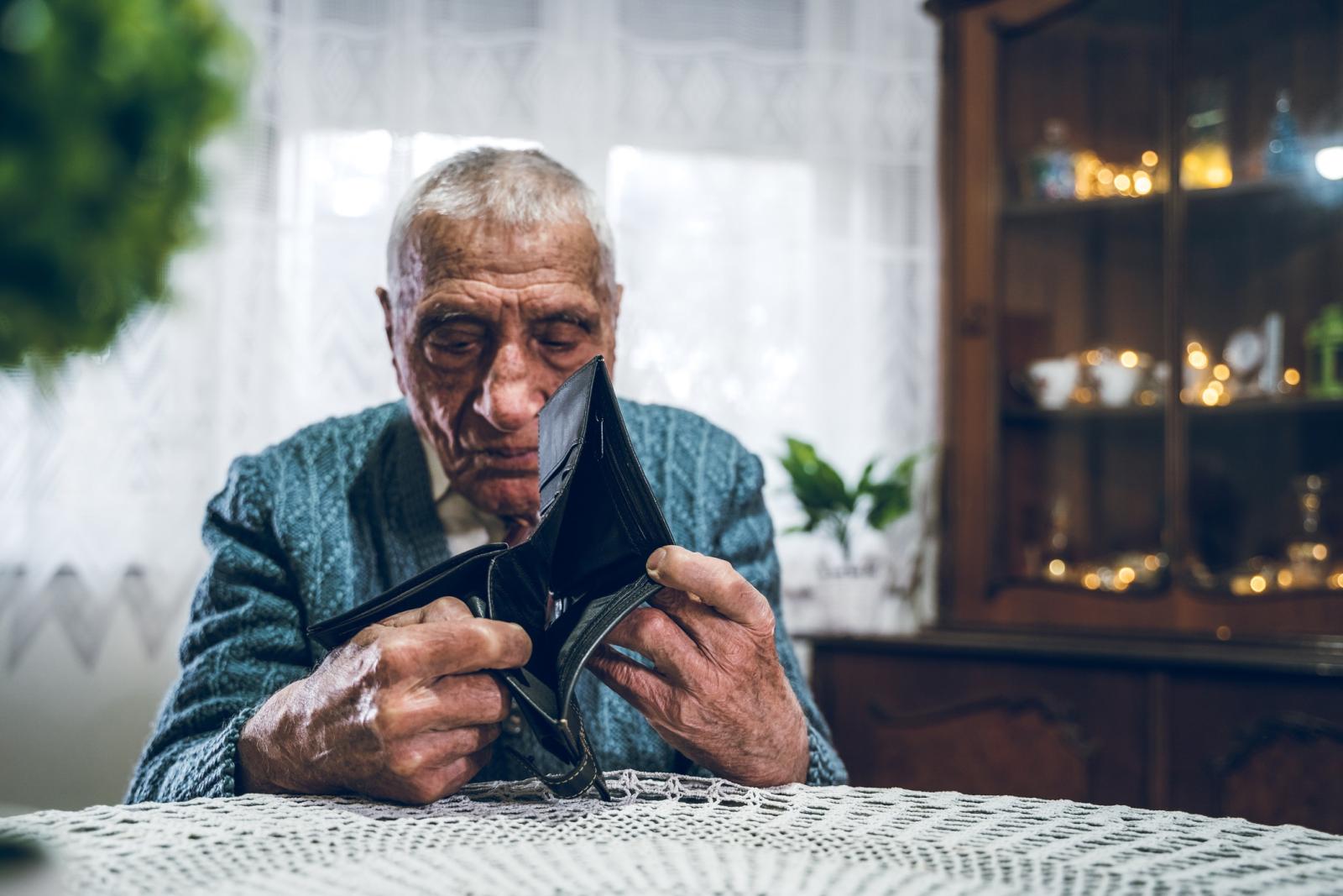 Een bejaarde man zit aan tafel en heeft een portemonnee in zijn handen. Er zit geen geld in de portemonnee.
