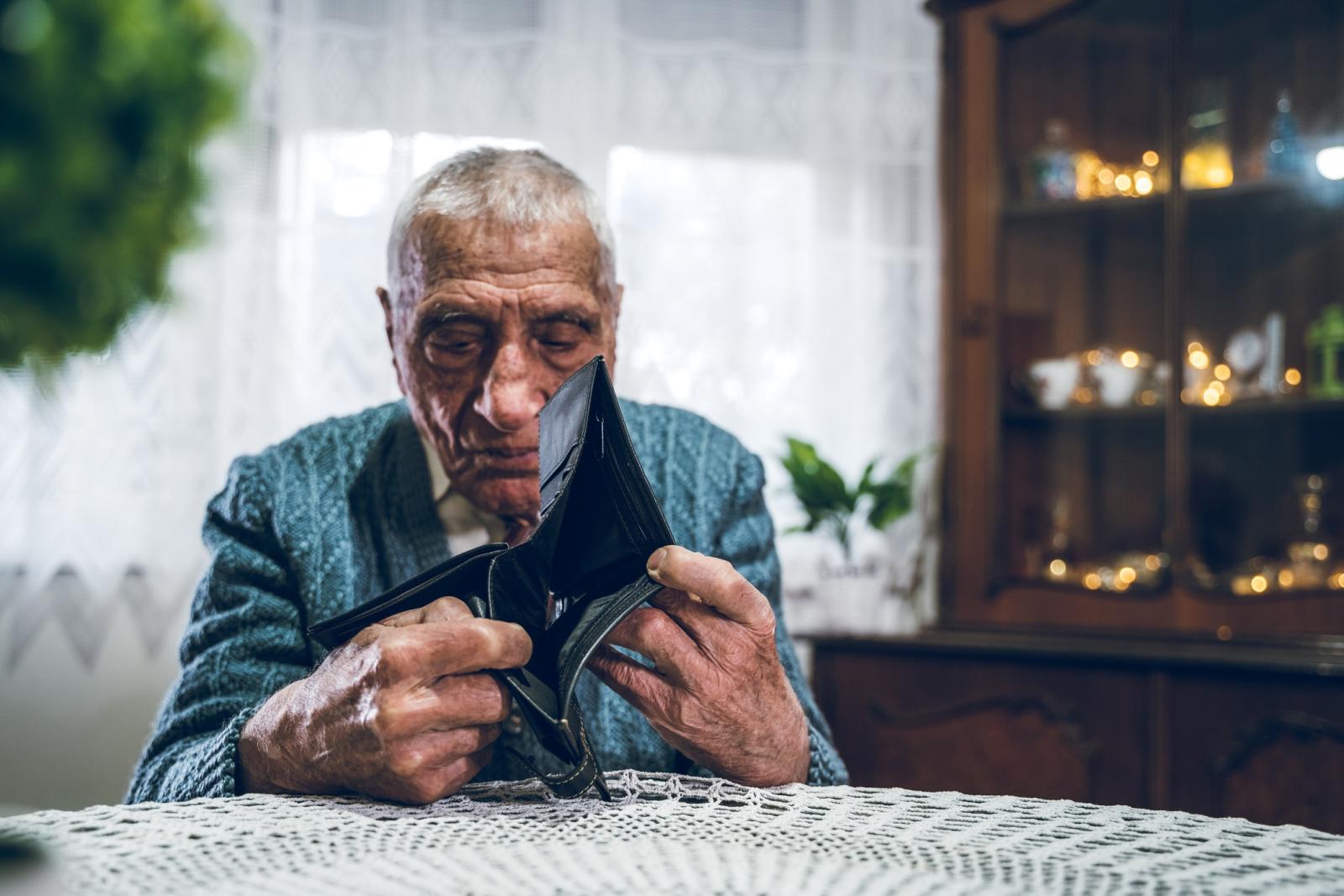 Een bejaarde man zit aan tafel en heeft een portemonnee in zijn handen. Er zit geen geld in de portemonnee. Een bejaarde man zit aan tafel en heeft een portemonnee in zijn handen. Er zit geen geld in de portemonnee.