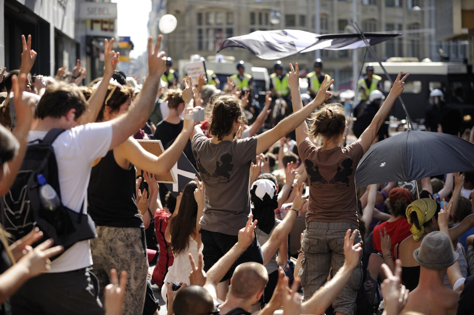 Je ziet een groep mensen demonstreren. Ze zijn van achteren gefotografeerd en houden allemaal hun armen omhoog en steken twee vingers op.