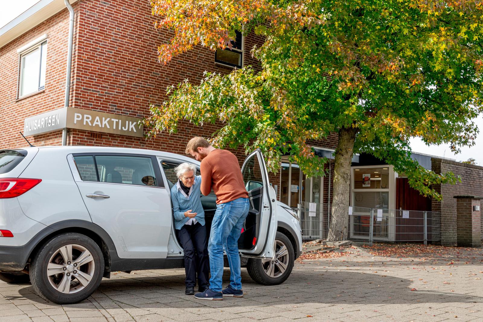 Een oudere dame wordt uit de auto geholpen door haar mantelzorger. Ze staan voor de huisartsenpraktijk. Een oudere dame wordt uit de auto geholpen door haar mantelzorger. Ze staan voor de huisartsenpraktijk.